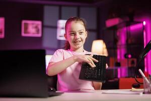 Child influencer filming wireless mechanical keyboard review in studio, showing capabilities and specifications. Little girl clicking keys on gaming computer peripheral, testing durability photo