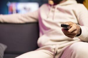 Man zapping through TV channels using remote, relaxing on couch in living room used as home theatre, close up shot. BIPOC person sitting on sofa, using television set to stream entertainment media photo