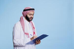 Muslim man dressed in traditional clothes holding clipboard and writing with pen. Person dressed in arabic thobe and headscarf taking notes in checklist while posing in studio photo