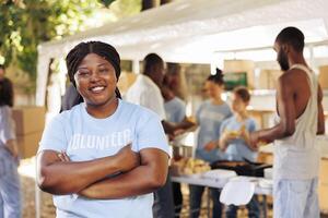 entusiasta negro mujer vistiendo azul camiseta con brazos cruzado mira a cámara. retrato Disparo de africano americano hembra voluntario, Listo a proporcionar humanitario ayuda a pobre, necesitado y Menos afortunado. foto