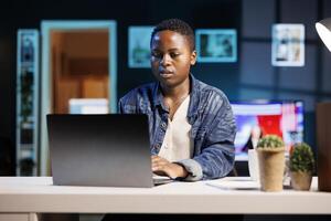 Busy black female freelancer sitting in her modern living room office, working on personal computer. African american college student typing and reviewing information on digital laptop. photo