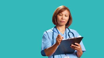 Close up shot of proficient healthcare expert writing appointment data of incoming patients on clipboard while at work. BIPOC nurse isolated over blue studio background finishing paperwork photo