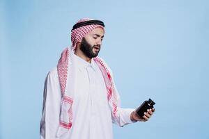 Muslim man dressed in cultural clothes holding medication and reading instruction on bottle. Person wearing traditional arabian robe and headscarf checking vitamins package photo