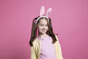 joven confidente niña con coletas y conejito orejas posando en cámara, sensación contento y alegre acerca de Pascua de Resurrección celebracion. pequeño niño siendo adorable terminado rosado fondo, festivo primavera día festivo. foto