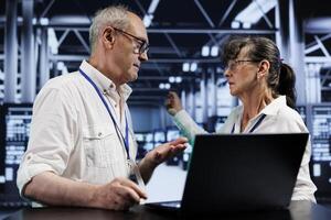 Senescent experts discussing how to best prevent strain on server hub systems caused by AI applications driving up power usage. Precise repairmen maintenancing rackmounts doing intense computations photo
