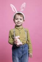 Joyful little boy holding a fluffy stuffed rabbit in front of on camera, wearing bunny ears and feeling excited about easter celebration. Small kid smiling and being happy against pink backdrop. photo