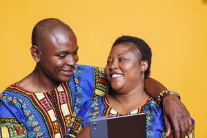 Cheerful smiling black couple wearing ethnic clothes hugging while enjoying online activity and using digital tablet. Happy pair in relationship embracing and scrolling social media feed together photo