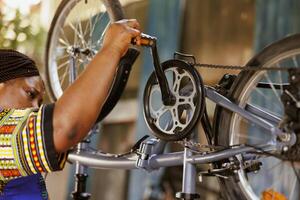 apasionado amante de los deportes negro mujer diligentemente inspeccionando y reparando bicicleta pedales y cadenas activo africano americano hembra ciclista haciendo anual ajustes a moderno bicicleta. foto