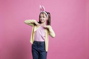 Adorable kid wearing bunny ears for easter photoshoot in studio, celebrating spring holiday over pink background. Cute little girl being playful and festive for traditional celebration. photo