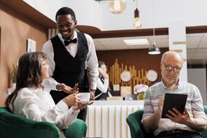 Elderly couple on hotel lobby sofa, using digital tablet and drinking coffee while employee ensures comfortable stay. Retired senior adults enjoy cup of tea and browsing internet in cozy lounge area. photo
