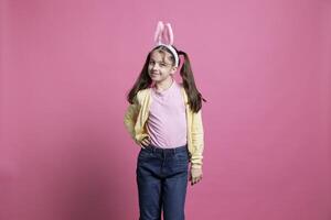 Little girl feeling happy and dancing in front of the camera, fooling around and acting silly against pink background. Young toddler wearing bunny ears and posing with confidence in studio. photo