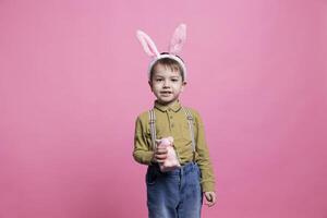 dulce alegre joven chico posando con un mullido Conejo juguete en cámara, vistiendo conejito orejas y celebrando Pascua de Resurrección fiesta festividad. pequeño niño siendo emocionado y alegre en contra rosado fondo. foto