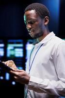 African american admin using laptop to review energy consumption across server rack parts. Professional making sure data center electronics temperature sensors are in perfect working condition photo