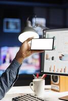 African american man holds device with white screen layout at his home desk, using funds analytics to solve business tasks remotely on computer. Freelancer works on web presentation, empty display. photo