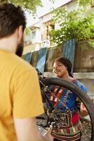 Sports-loving black woman checking and maintaining bicycle for yearly summer cycling. Active young caucasian man helps healthy energetic african american woman mend damaged bike wheel outside. photo