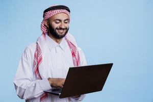 sonriente hombre vestido en musulmán thobe y Pañuelo mensajes de texto en línea mientras en pie y participación ordenador portátil. alegre árabe persona vistiendo tradicional atuendo mecanografía en portátil computadora teclado foto