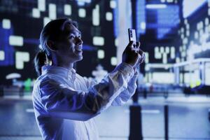 BIPOC man walking around city at night, using mobile phone to take photos. Asian citizen using cellphone to take pictures while strolling outside on empty streets illuminated by lamps photo