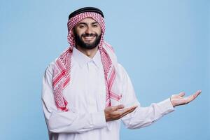 Muslim man in traditional robe confidently pointing to left side with hands studio portrait. Arab showcasing product advertisement and looking at camera with cheerful expression photo