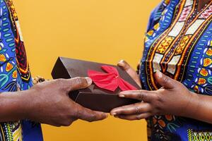 Black couple exchanging anniversary gift, holding in hands wrapped box with bow ribbon closeup. African american man and woman sharing festive present, receiving box close view photo