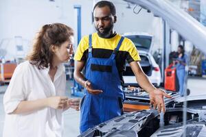 Repairman at auto repair shop conducts routine vehicle checkup, informing customer about needed engine fix. BIPOC garage worker chatting with customer after finishing car maintenance photo