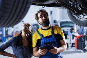 Hardworking specialist helping customer with car checkup in auto repair shop. Skilled garage worker looking over automobile parts with woman, repairing her broken vehicle wheels during inspection photo