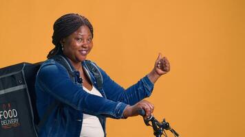 On yellow background, african american woman carrying food delivery bag on her back. Female bike messenger on bicycle pointing to isolated copyspace backdrop template with hand motions. photo