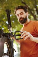 Sports-loving dedicated man repairing bicycle parts using professional tools in home yard. Healthy caucasian male cyclist assessing and mending damaged bicycle as summer activity. photo