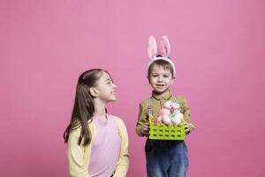 Cute brother and sister showing festive painted eggs and rabbit toy in a basket, enjoying easter celebration holiday. Little kids siblings posing against pink background, handmade decorations. photo