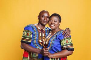 Happy african american romantic couple hugging and looking at camera with joyful expression. Smiling man and woman wearing ethnic clothes hugging and posing for portrait on orange background photo
