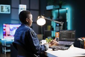 African American freelancer working at a desk focused on her laptop that is displaying code and data. Black woman with wireless computer, programs and manages the software, ensuring cyber security. photo