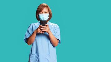 Happy relaxed nurse chatting with friends online while taking hospital shift break. Smiling woman typing message on phone, wearing medical scrubs, isolated over blue studio background photo