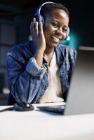 Black woman joyfully using a laptop and wireless headphones. African american female influencer multitasks, attending an online meeting while listening to music in her headset. photo