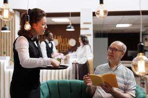 In lounge area, young waitress serving a cup of tea to elderly male tourist seated on couch with a book. Retired senior man sitting on cozy sofa and being attended by female employee in hotel lobby. photo