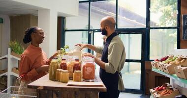 comerciante regalos abultar productos en frascos, recomendando tipo de bio orgánico pasta a mujer cliente en local tienda de comestibles almacenar. joven adulto demostración eco artículos en reciclable contenedores trípode disparo. foto