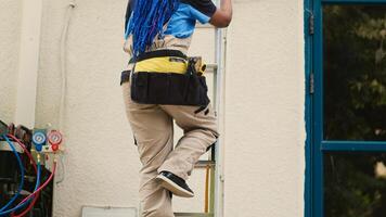 African american specialist in professional uniform with technical equipment climbing folding ladder to do maintenance on rooftop air conditioner. Trained engineer commissioned to checkup on condenser photo