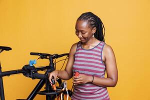 joven africano americano dama preparando esencial herramientas para zurcidura dañado bicicleta. deportivo negro mujer agarrando varios especializado equipo para reparando roto bicicleta en contra un aislado antecedentes. foto