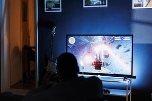 Young adult enjoying time playing shooter games on tv, plugging his console to television in the living room and spending game session at night. Man engaging in first person shooting contest. photo