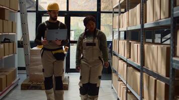 Employee and african american coworker walking through warehouse aisles checking labels on cardboard boxes sitting on shelves, inputing updated products info on online shopping website photo