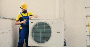 experto Wireman comenzando trabajo en mal funcionamiento aire acondicionador, utilizando mano perforar a tomar aparte condensador bobina panel. competente mecánico desmantelamiento hvac sistema a cheque para defectuoso alambres foto