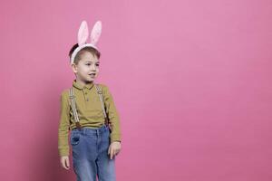 Little boy being cute with bunny ears in front of the camera, feeling excited about easter celebration event and receiving presents. Cheerful small kid feeling joyful against pink background. photo