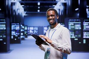 Portrait of african american administrator in front of server clusters in high performance computing data center. Technician monitoring energy consumption across systems, ensuring optimal operations photo