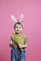 Young cute toddler playing around with a toy in front of camera, feeling happy while he wears fluffy bunny ears. Joyful small child posing against pink background, innocent kid. photo