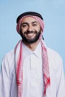 Cheerful muslim man dressed in islamic white thobe and checkered ghutra headdress studio portrait. Happy arab person standing and looking at camera with carefree expression photo