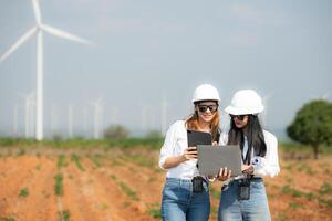 Two asian engineer women holding laptop and working in wind turbine farm with blue sky background photo