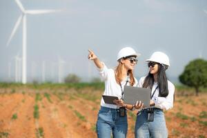 Two asian engineer women holding laptop and working in wind turbine farm with blue sky background photo