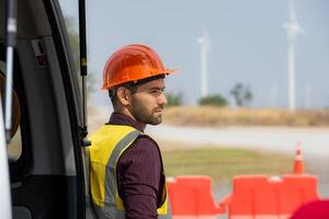 Portrait of a young male engineer standing behind a car in wind turbine farm with blue sky background photo