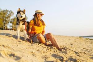 Woman in hat and sunglasses sits with her dog on the beach and enjoying sunset photo