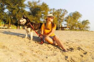 Woman in hat and sunglasses sits with her dog on the beach and enjoying sunset photo