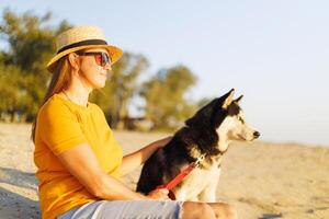 Woman in hat and sunglasses sits with her dog on the beach and enjoying sunset photo