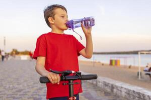 A boy drinks water from plastic bottle, standing with scooter photo